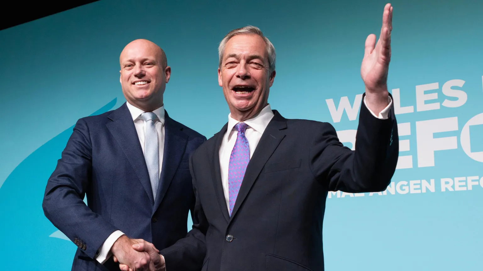 Dan Thomas and Nigel Farage standing front of a Reform UK logo. Farage has one hand in the air waving to people.