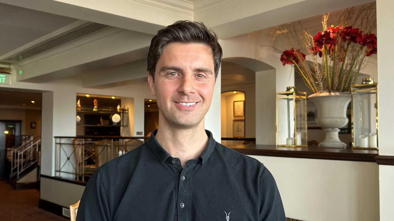 Matt Seymour, a man with short brown hair, is wearing a dark collared shirt. He is pictured in a hotel lobby. To the right of the image is a large vase of red flowers.