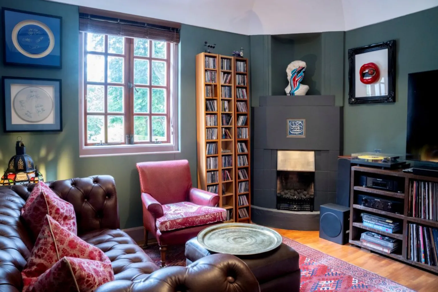 IWC Media Inside Art Deco Villa. The room has dark green walls, brown leather sofas and pink soft furnishings. A modern fire place is in the corner and there is a CD rack on the wall. Paintings hang on the walls and there is a window on the left.