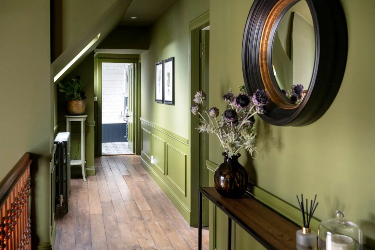 IWC Media Inside The Schoolhouse. The walls in the hall are olive green with a wooden floor. A large round mirror hangs on the wall and there is a vase on the sideboard.