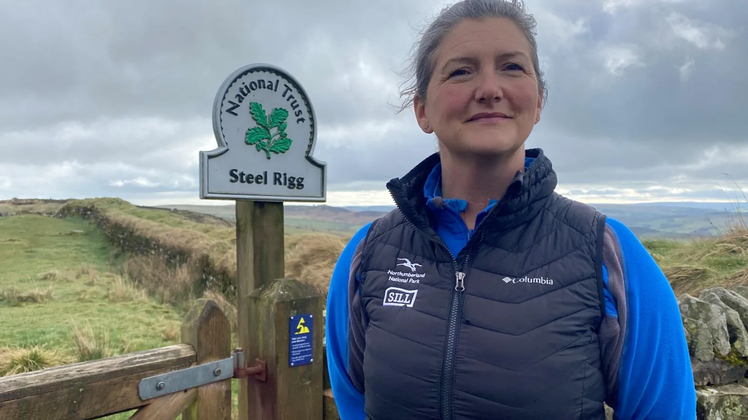 Margaret Anderson is a women in her 30s with blonde hair tied back. She is wearing a blue top with sleeves and a sleeveless darker blue jacket with the northumberland National park logo on it. Behind her is a stretch of Hadrian's Wall 