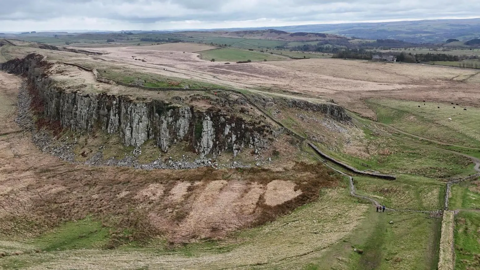 An aerial view of a stretch of Hadrian's Wall which stretches out into the distance along a geological structure called a sill which rises steeply out of the group meaning the wall sits on a cliff edge. 