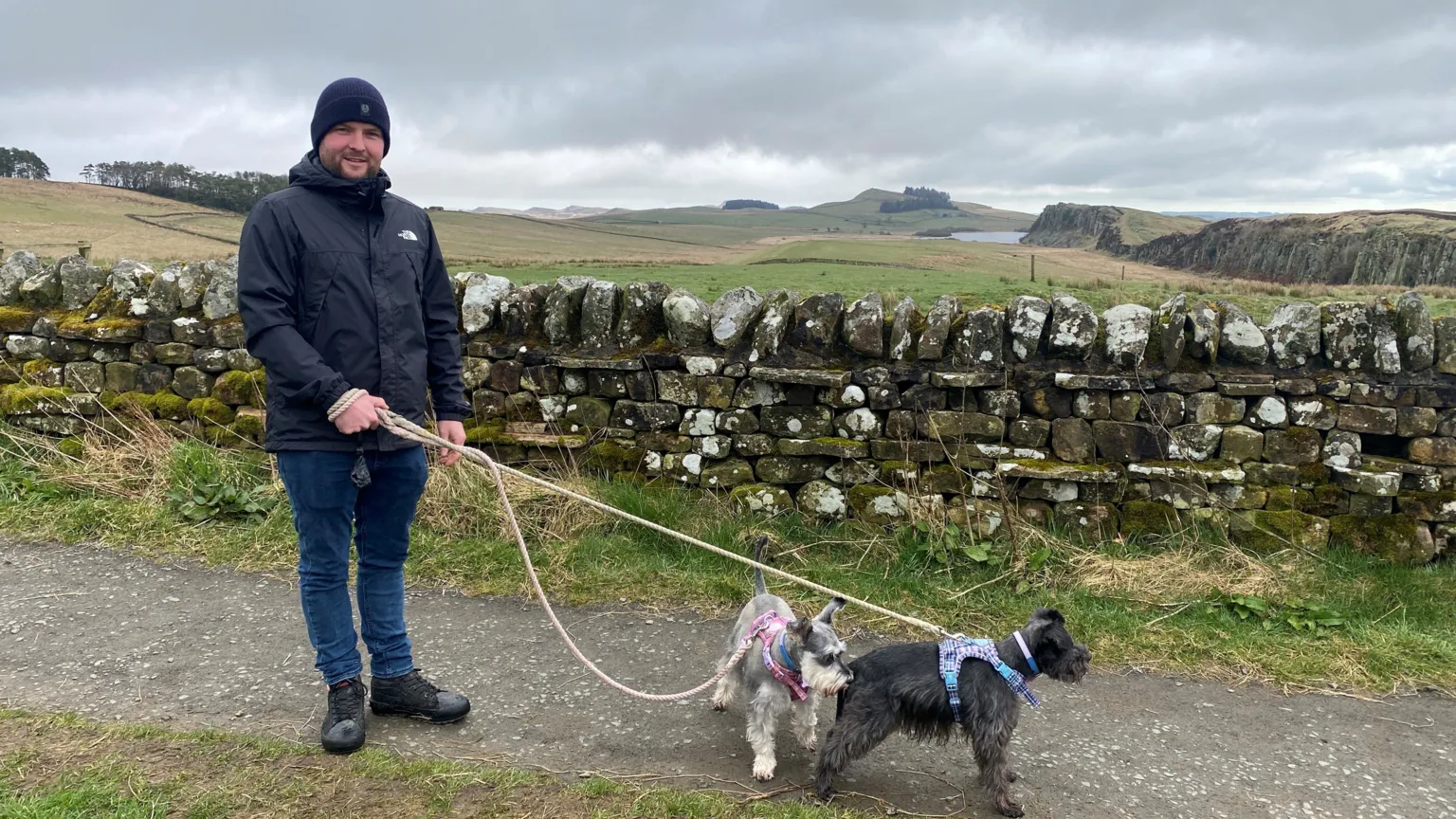 Taylor Hughes is a man in his thirties wearing a blue beanie, blue jeans and a dark blue top and black boots. He has two small dogs on leads. He is standing in front of a wall with a stretch of Hadrian's Wall visible
