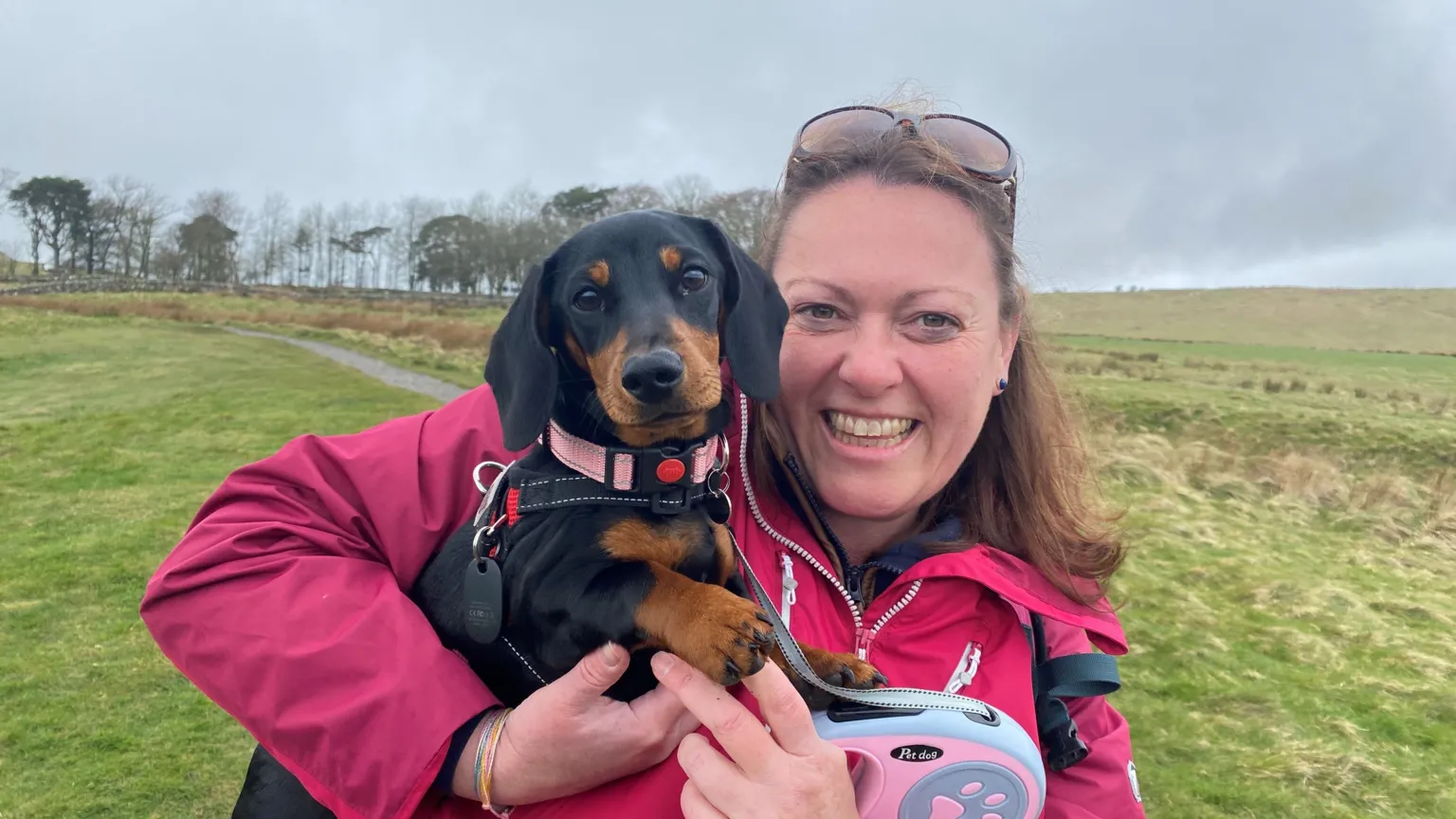 Emma Harrison is a young woman holding a dachshund in her arms. She is wearing a pink anorak and behind her is a long stretch of hillside with a path and a row of trees at the top 