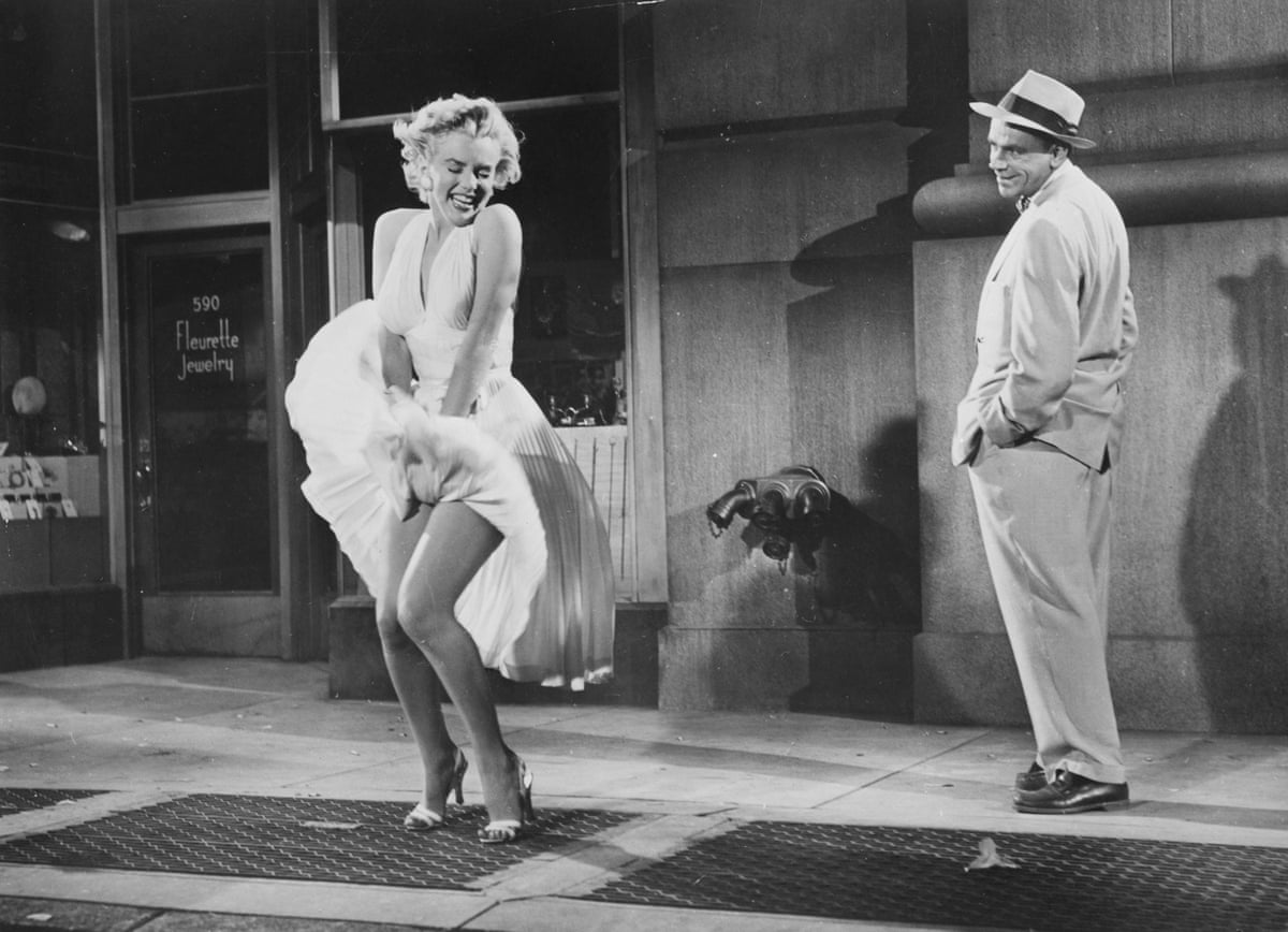 A black and white film still of Monroe in a white dress, standing next to a make actor