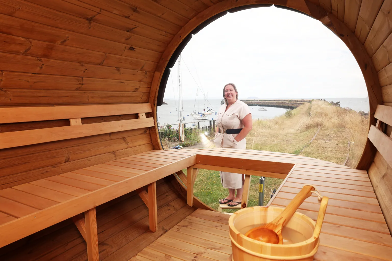 Môr A Sawna The image shows the inside of a wooden style sauna, Kathryn Donavan, the owner of the sauna is stood just outside. She's wearing a beige dress with a black belt and a pair of black flip flops. She has short blonde hair.