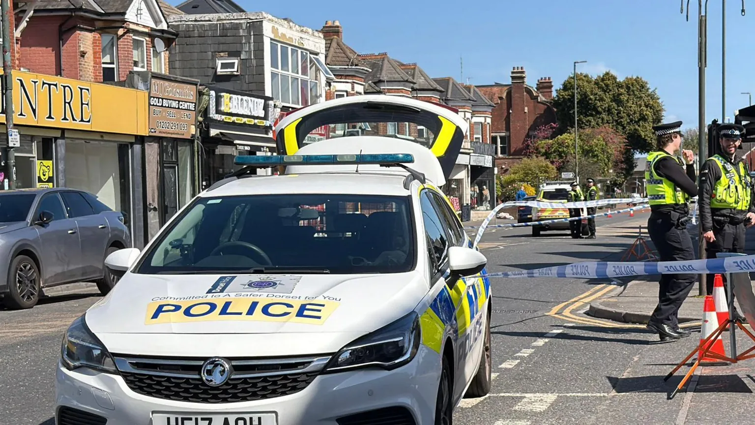 Police car parked with front facing the camera, police tape is across a road where two police officers are standing next to a road closed sign. 
