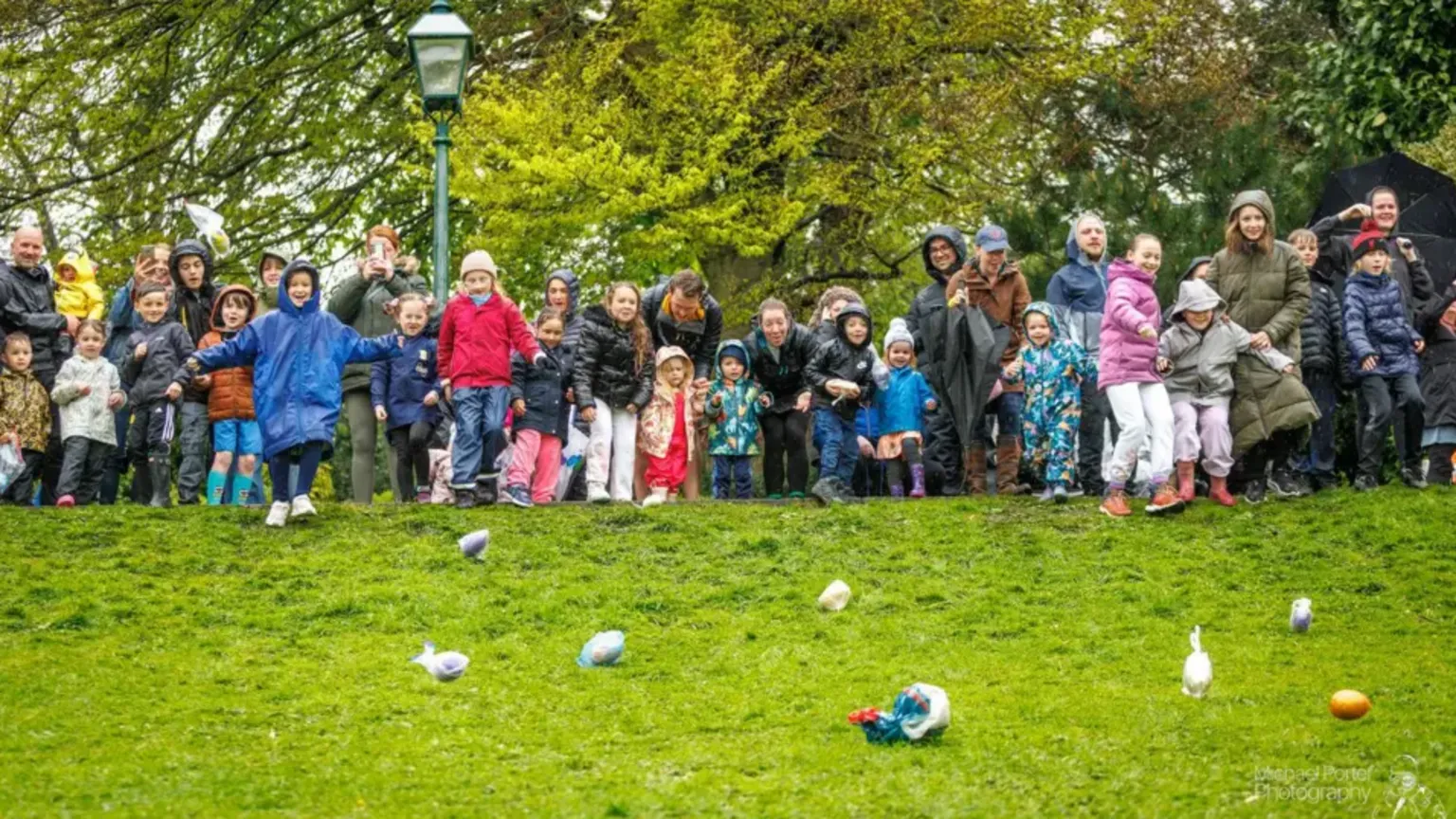Micheal Porter Photography A group of people wearing coats and winter clothing stand at the top of a grassy hill, with large, designed eggs rolling down it. Some of the eggs are in plastic bags. There are leafy trees and a lamppost in the background.