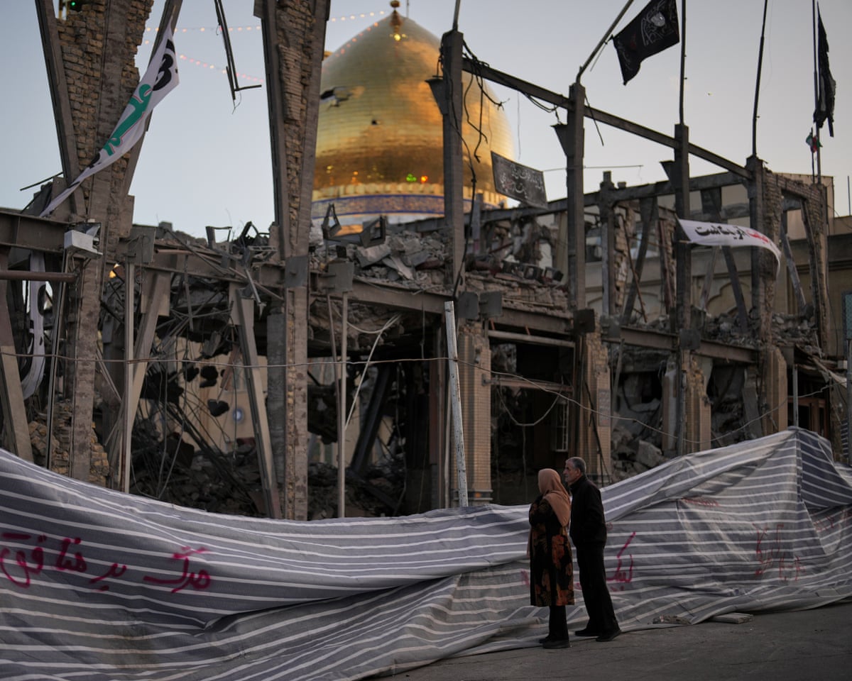 People look at a destroyed building within the Grand Hosseiniyeh, with the mosque visible in the background, after it was hit in airstrikes.