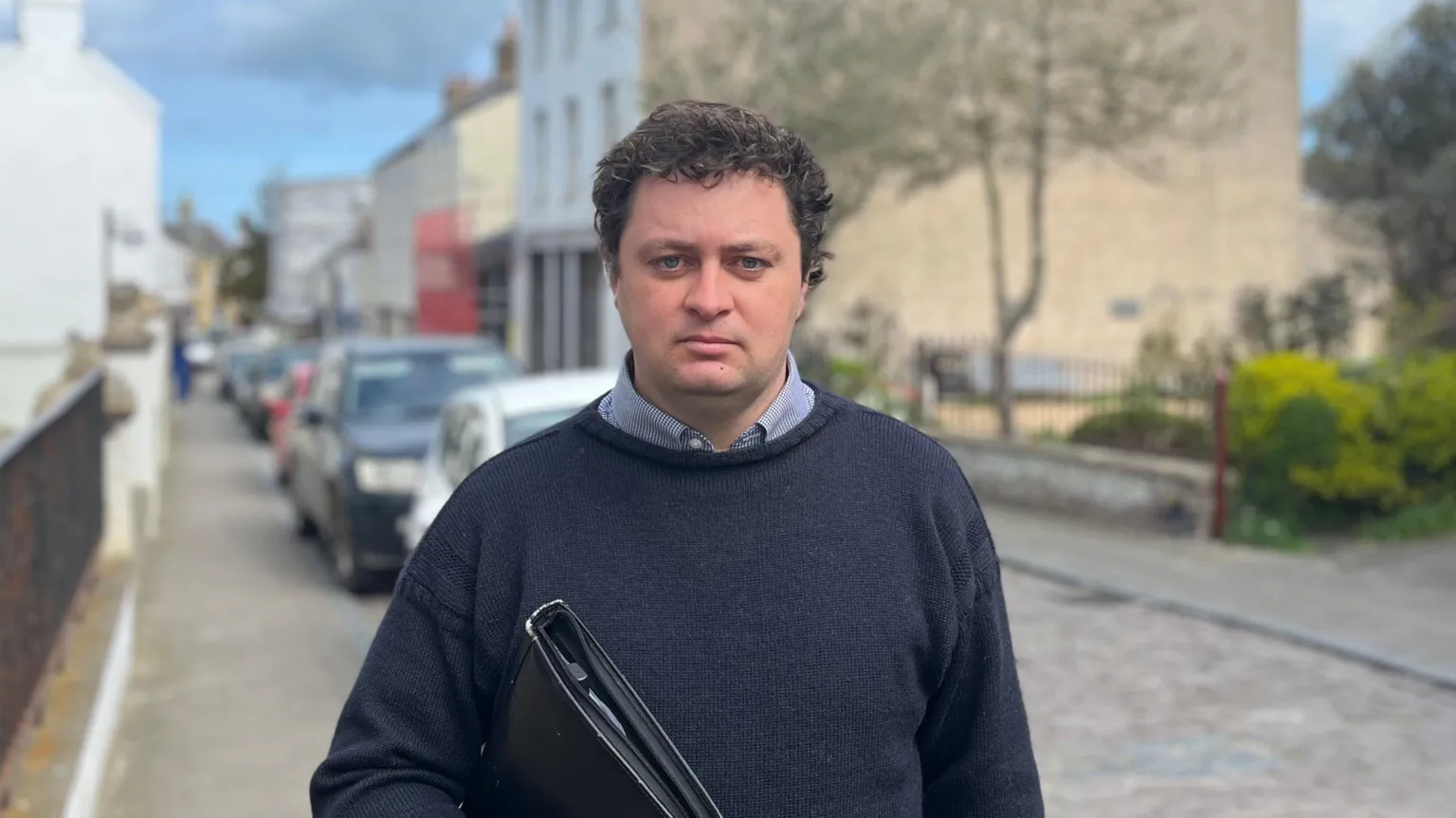 Alex Snowdon facing the camera, stood on Victoria street in Alderney. He is weaing a Navy Guernsey Jumper and holding a binder.