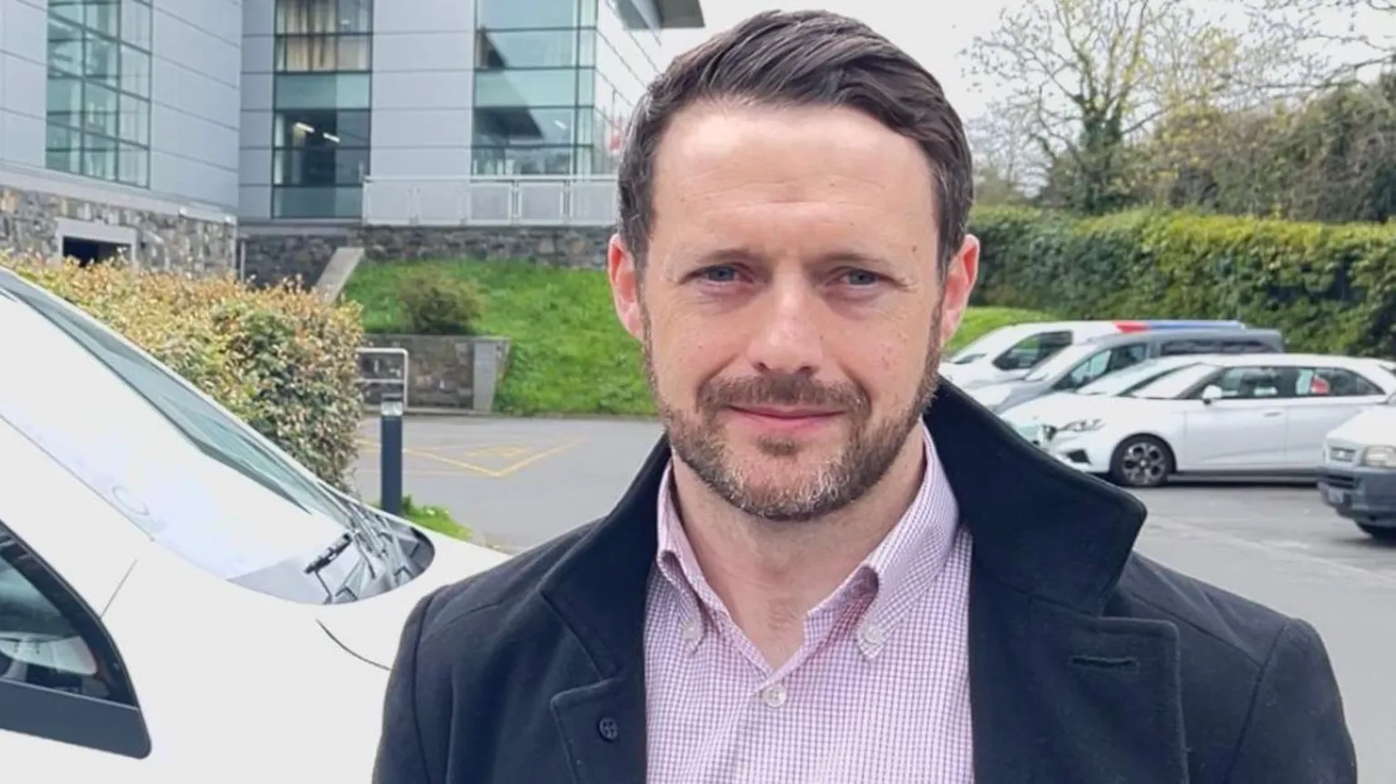 Niall Mc Swiggan facing the camera in front of parked cars. He is wearing a collared jacket and a button up shirt. He has short dark hair and a short beard.
