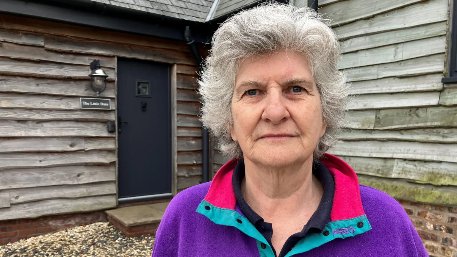 An woman with short grey hair and a purple fleece standing outside the annexe attached to her home. The building have wooden cladding.