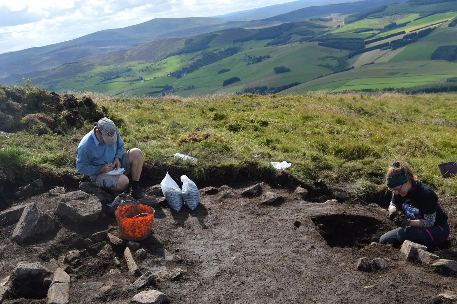 University of Aberdeen Two archaeologists sit on bare soil in an excavated area on the hill. Behind them is a landscape of rolling green, farmland.