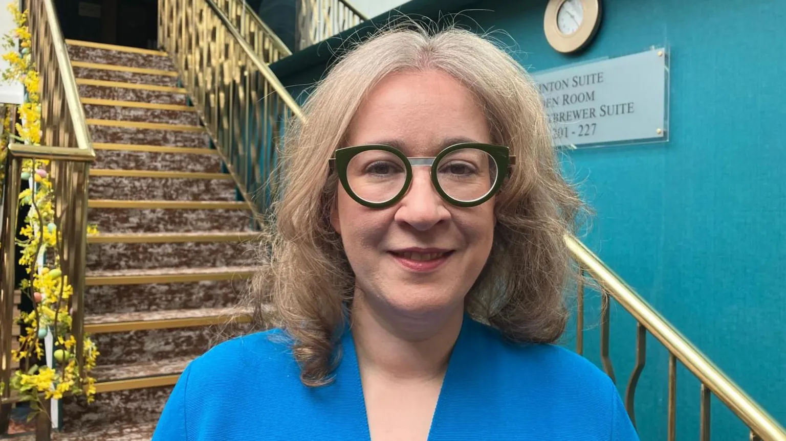 Selina Horshi has shoulder-length blonde hair and round glasses and is standing in front of a staircase which has a brown coloured carpet and brass bannisters. She is wearing a dark blue top. Over her left shoulder a teal coloured wall can be seen.