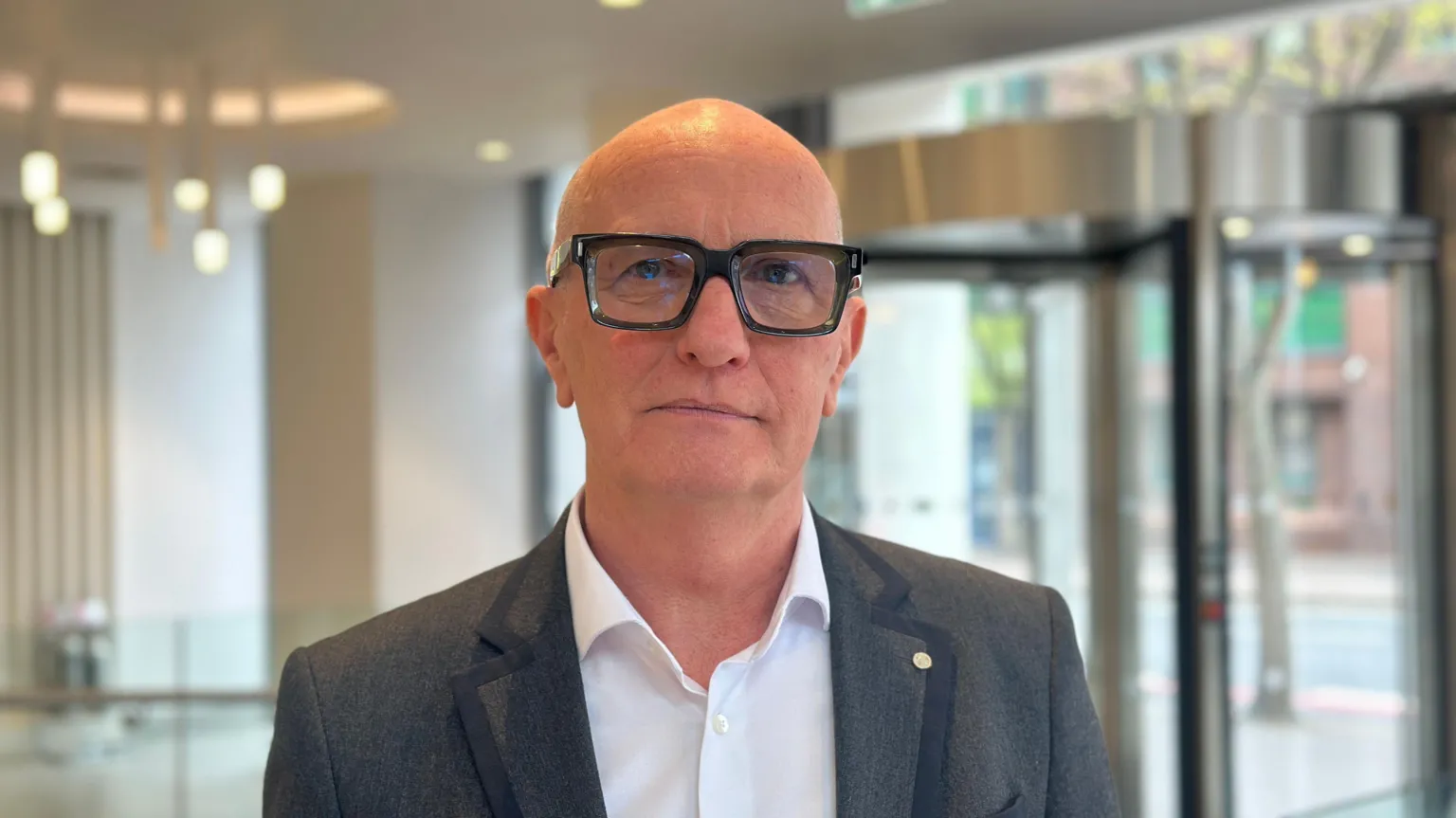 Colin Neill is a bald man with black thick rimmed glasses. He is wearing a grey suit jacket and a white shirt which is open at the collar. He is standing in the lobby of a hotel - behind him revolving doors can be seen.