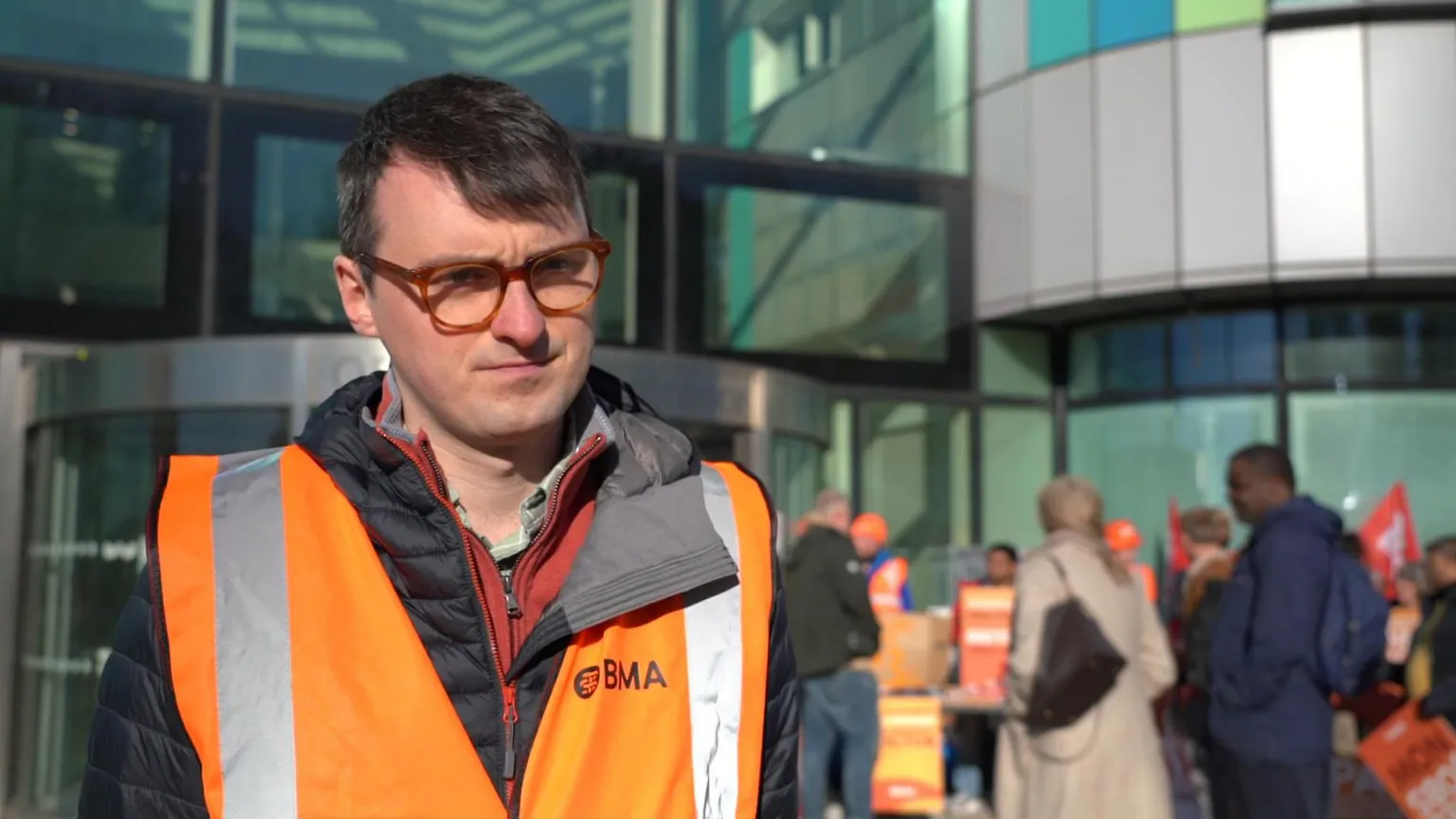 A man with dark hair in an orange hi-vis jacket and glasses is on the left of the photo. People near a building are blurred in the background on the right of the image.