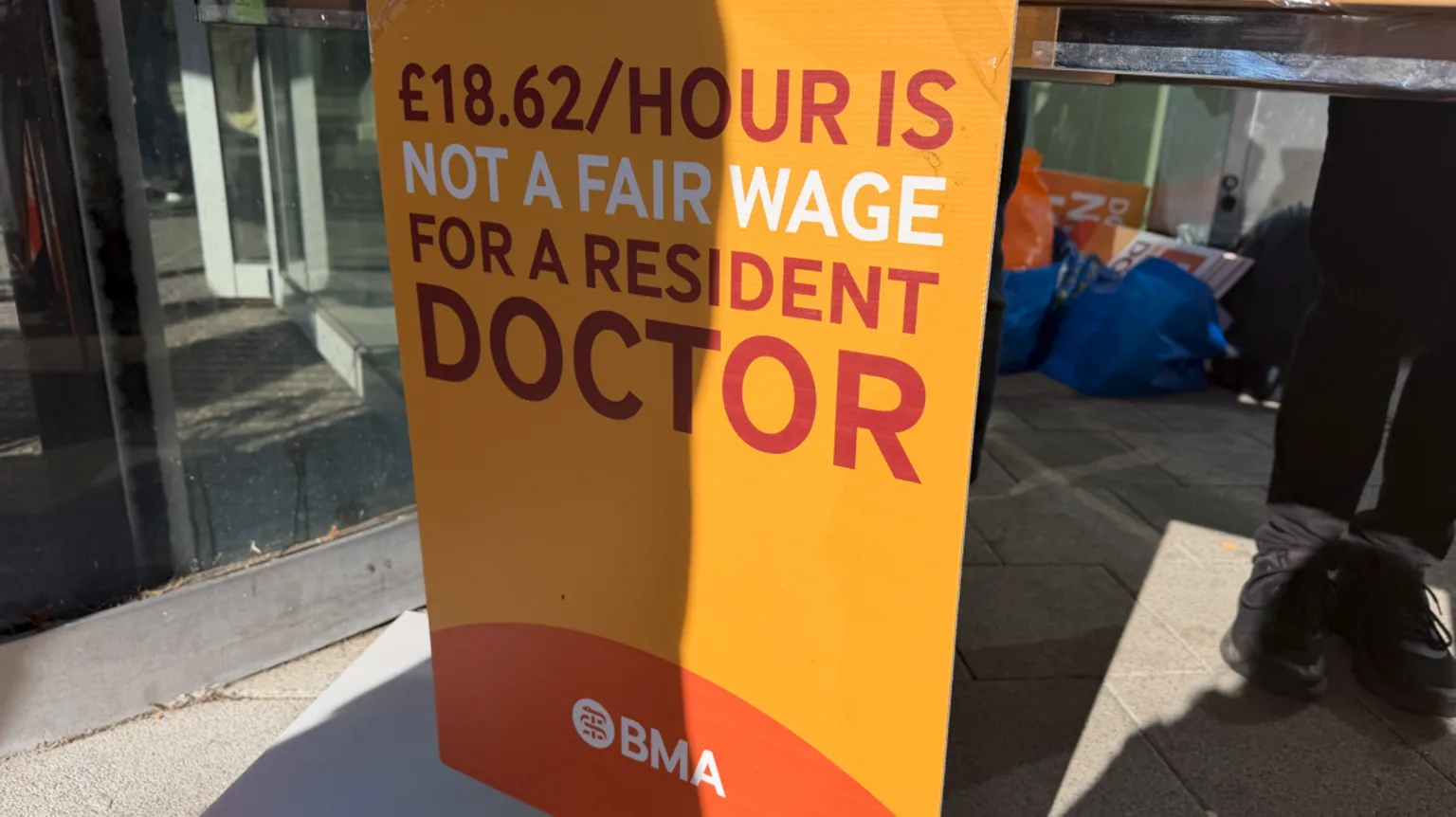A banner, which is yellow, white and orange, including £18.62/hour is not a fair wage for a resident doctor is prominent in the middle of the image.