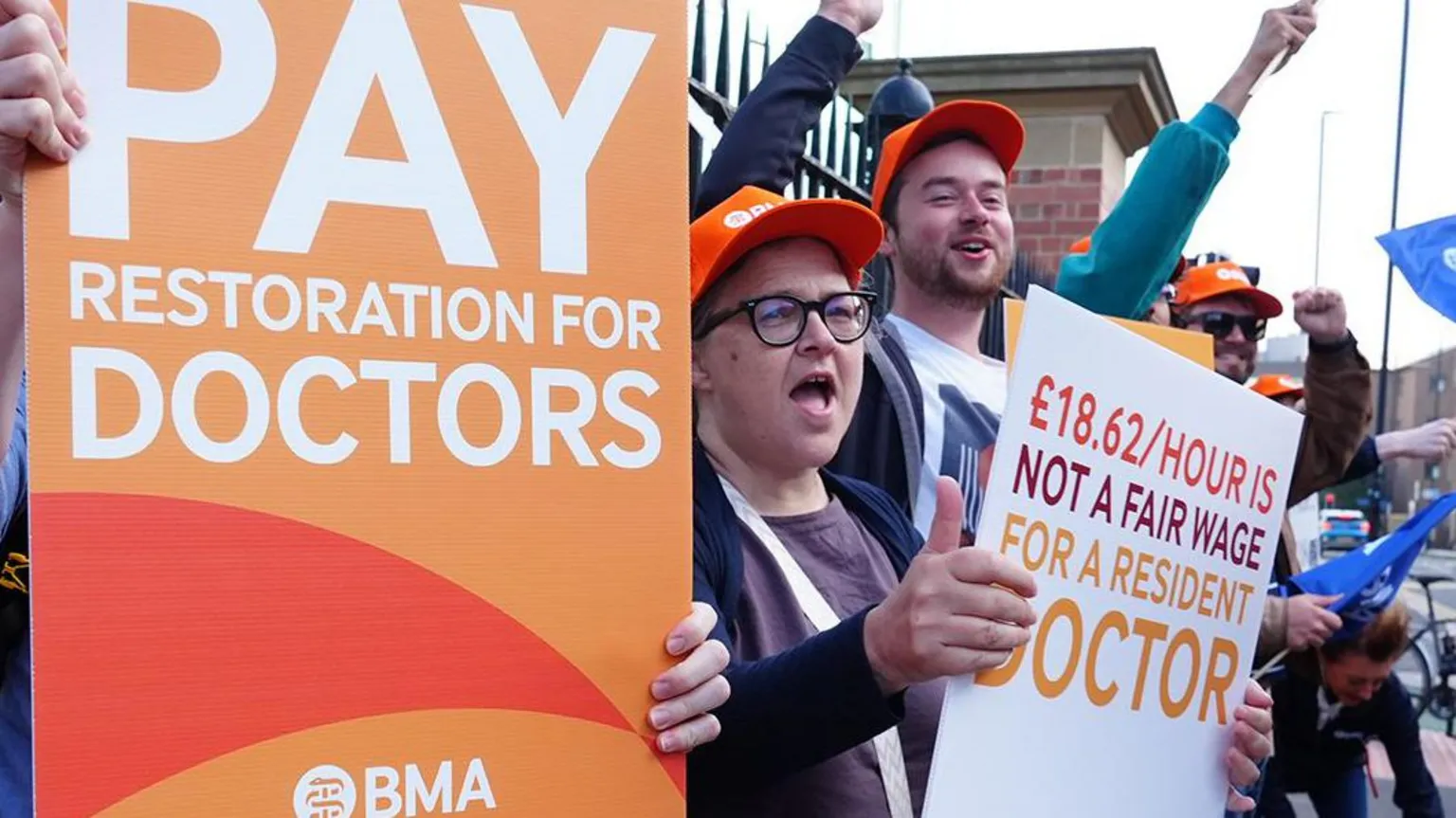 A group of doctors wearing orange BMA hats protest outside iron railings, chanting and raising their fists while holding placards reading “Pay restoration for doctors” and “£18.62/hour is not a fair wage for a resident doctor