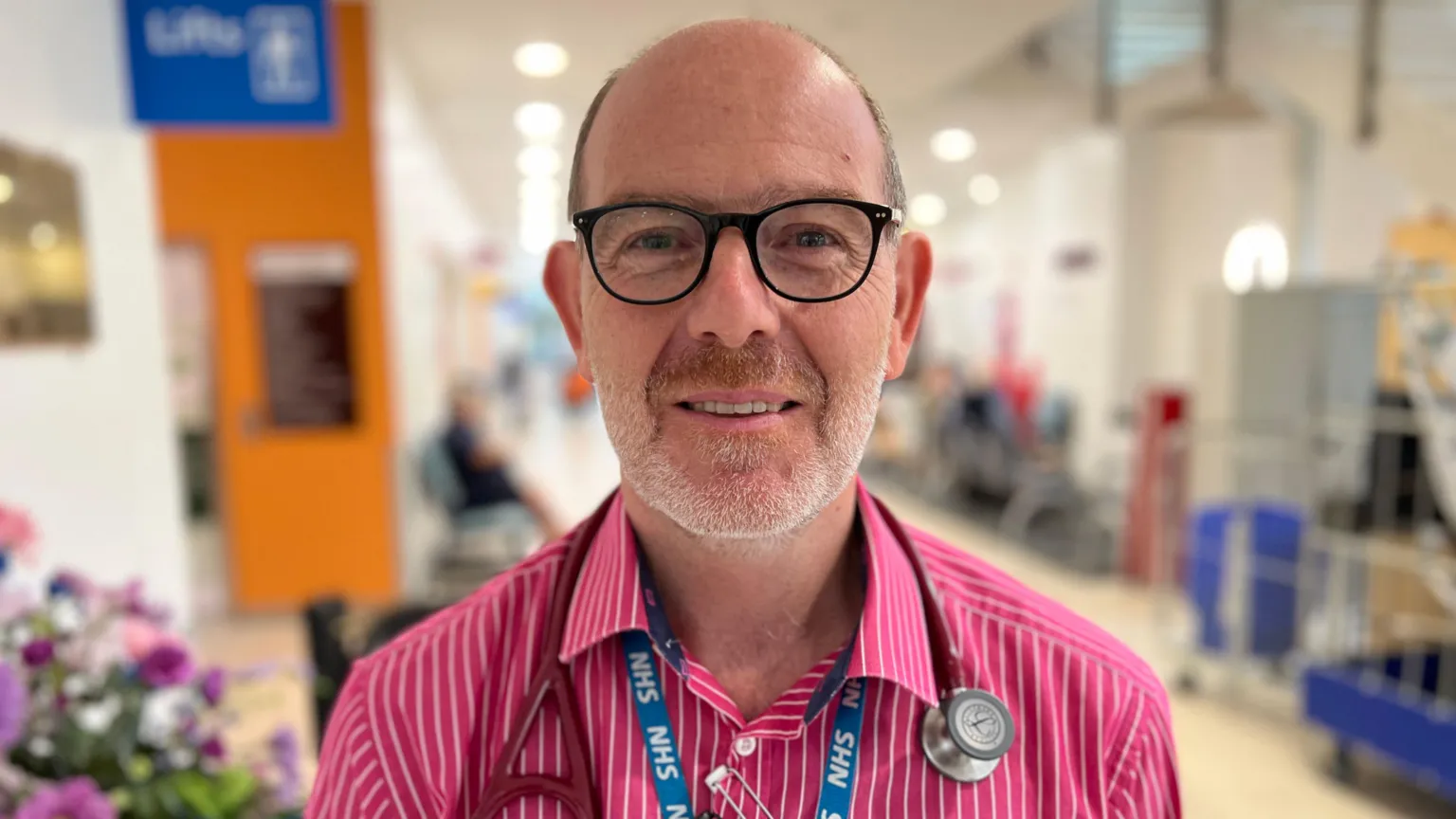 A middle-aged man with very short brown hair and a receding hairline stands smiling in a brightly lit hospital corridor. He is wearing black-rimmed glasses, a blue NHS lanyard, and a pink shirt with thin white stripes. There is a stethoscope around his neck.