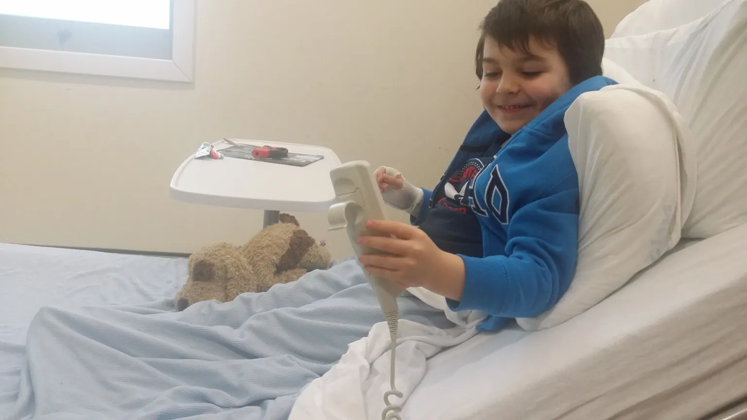 Cotter Family A young boy lying in a hospital bed, with a stuffed toy on the bed with him. He has dark hair and wearing a blue top.