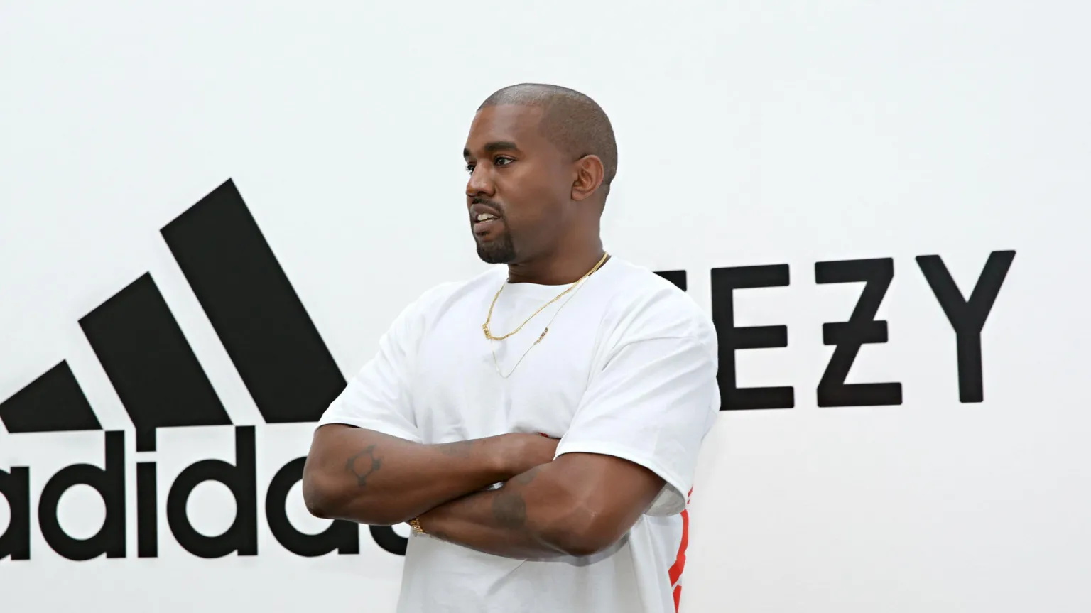 West in a white T-shirt with arms folded in front of Adidas/Yeezy sign