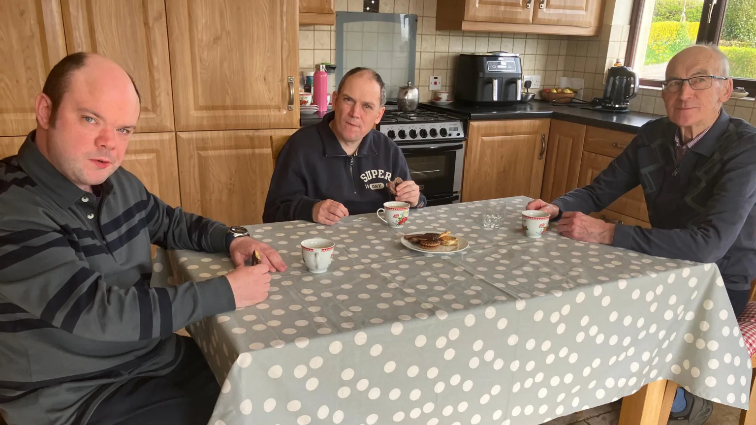 Alphie Lonergan sits at his kitchen table with his sons Ronan and Donal. Donal, in the middle, is wearing a navy quarter-zip top and has receding dark hair. Ronan is wearing a striped top and is bald on top with dark hair at the sides. The kitchen table has a grey/green tablecloth with white polka dots on it and there is a plate of biscuits in the middle. A mug is in front of each of the three men.