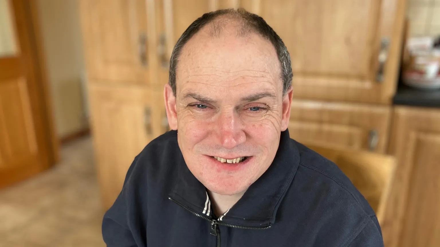 Donall Lonergan has receding dark hair and is wearing a navy quarter-zip top. He is smiling and behind him the oak cabinets of a kitchen can be seen.