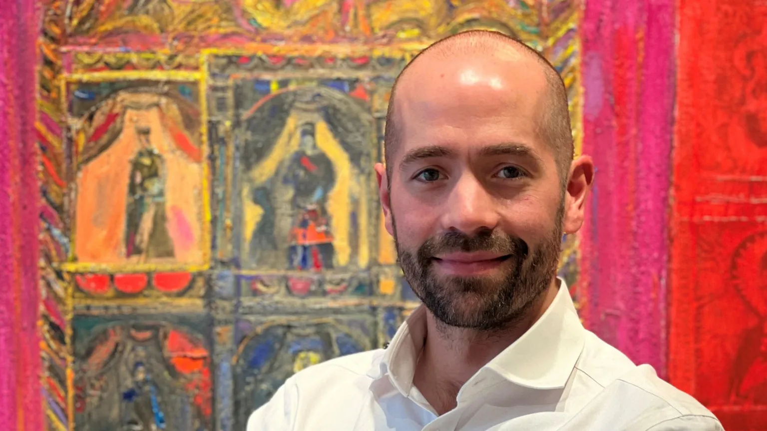 The Scottish Gallery Portrait of a man in a white shirt, smiling slightly while standing in front of a vivid, colourful painting with theatrical figures and bold red and gold tones.