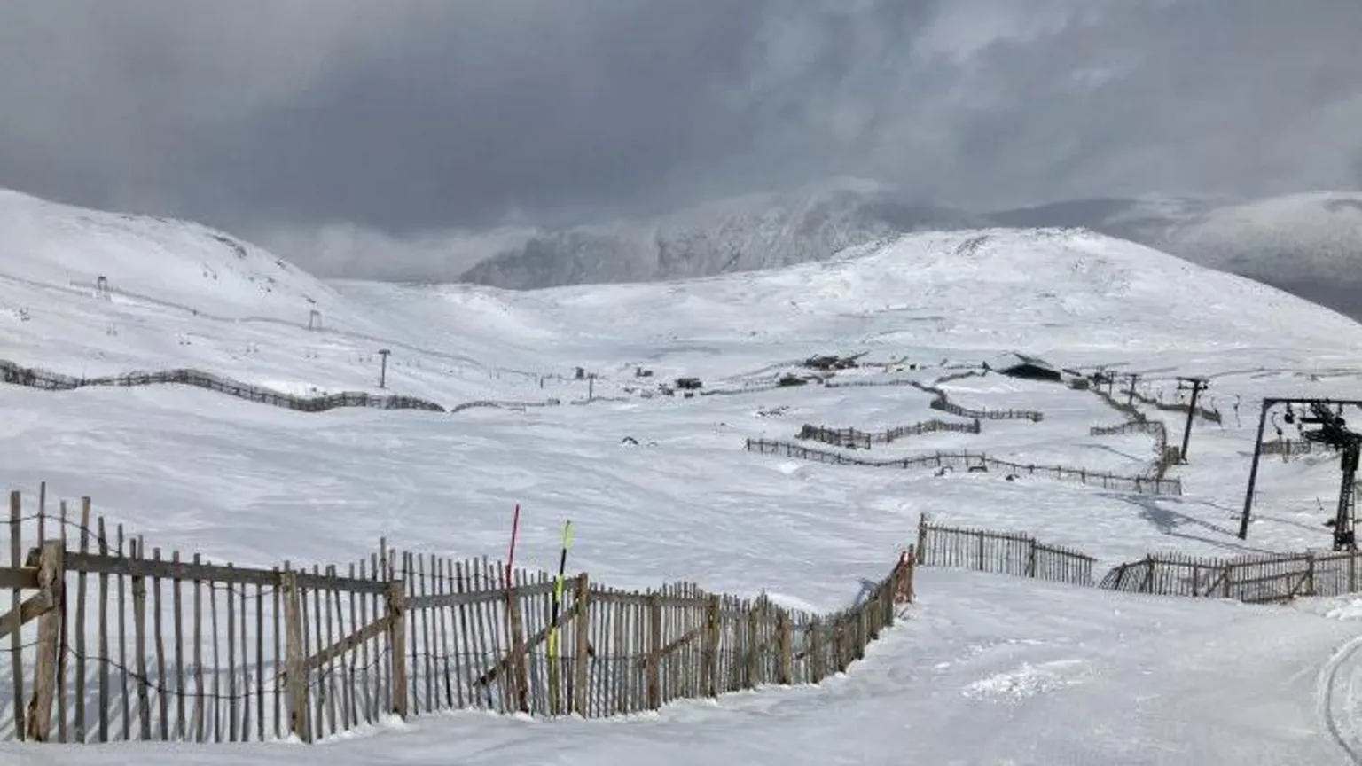 SAIS Glencoe Glencoe Mountain ski area is covered in snow. There is a snow fence and in the distance ski lift infrastructure. The sky above is grey and cloudy.