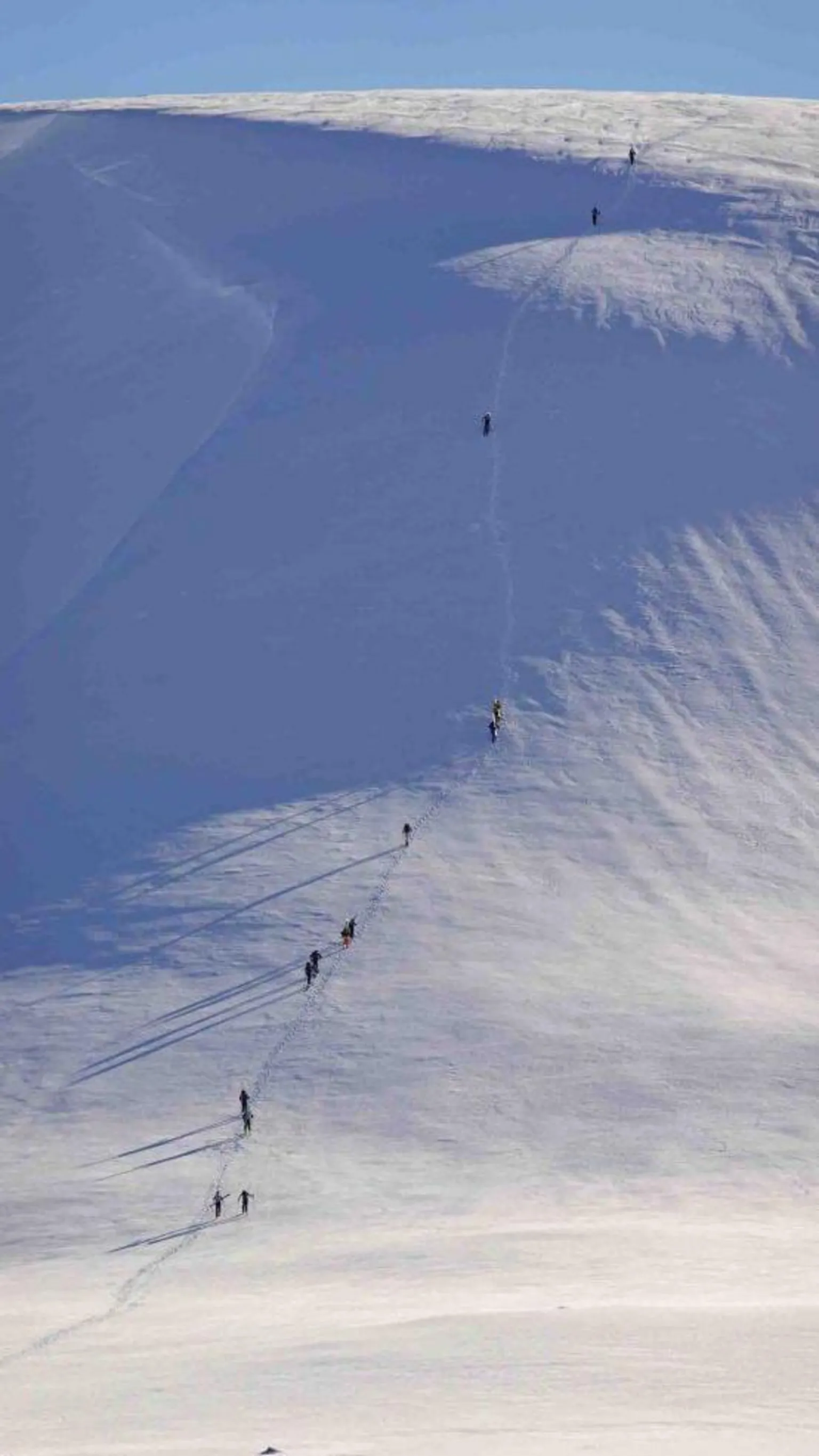 SAIS Southern Cairngorms Ski tourers walking up a snow covered slope at Glas Maol in the Southern Cairngorms. The skiers are walking in a line up a steep slope under a blue sky.