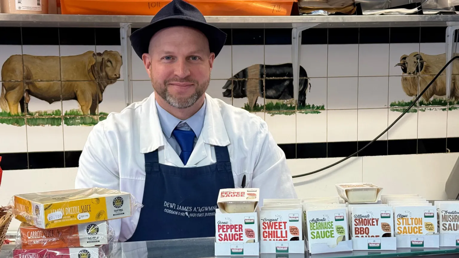 Butcher Dafydd Davies wears an apron and butchers coat and hat and stands next to his meat counter and a tiled wall with a pig, cow and sheep on the wall.