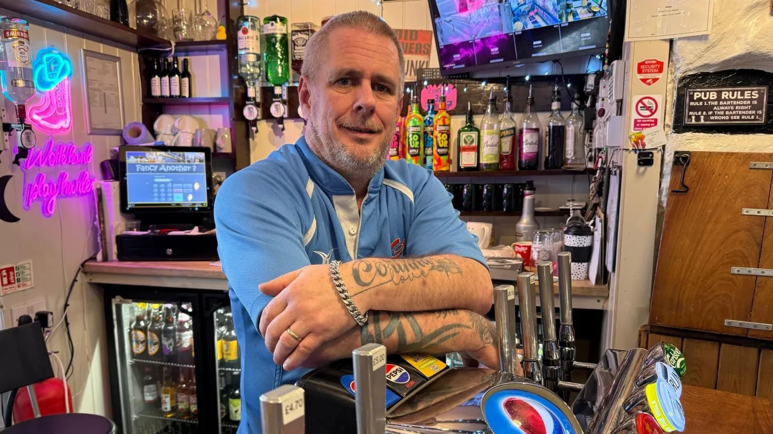 Landlord Sam Selby is pictured behind the bar leaning on pumps with tattooed arms and a short hair cut wearing a rugby jersey