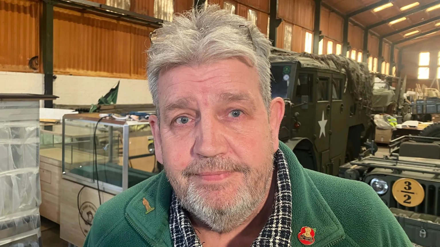 David is smiling at the camera. He has grey hair and beard. He is wearing a green fleece jacket with a poppy pin on his lapel. Behind him various vintage military vehicles can be seen sitting in a large warehouse.