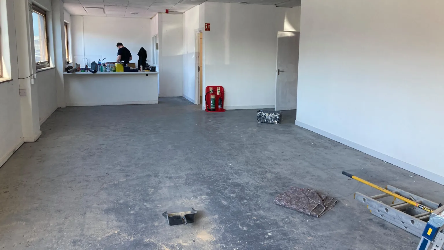 A large room with white walls. There are three square windows to the left. A builder wearing a black top is stood at the far end of the room, with his back to the camera. There is a ladder laying on the floor.