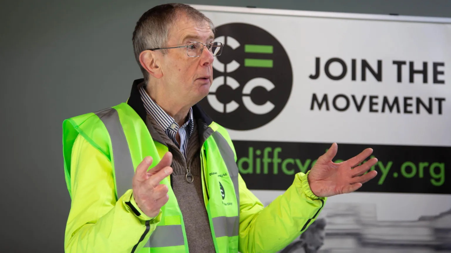 Eva Marloes Chris Roberts stands at an event with a viz vest and jacket open over his grey jumper. he is holding his hands up in the air at a he speaks in front of a campaign sign on the wall behind him that say join the movement. 