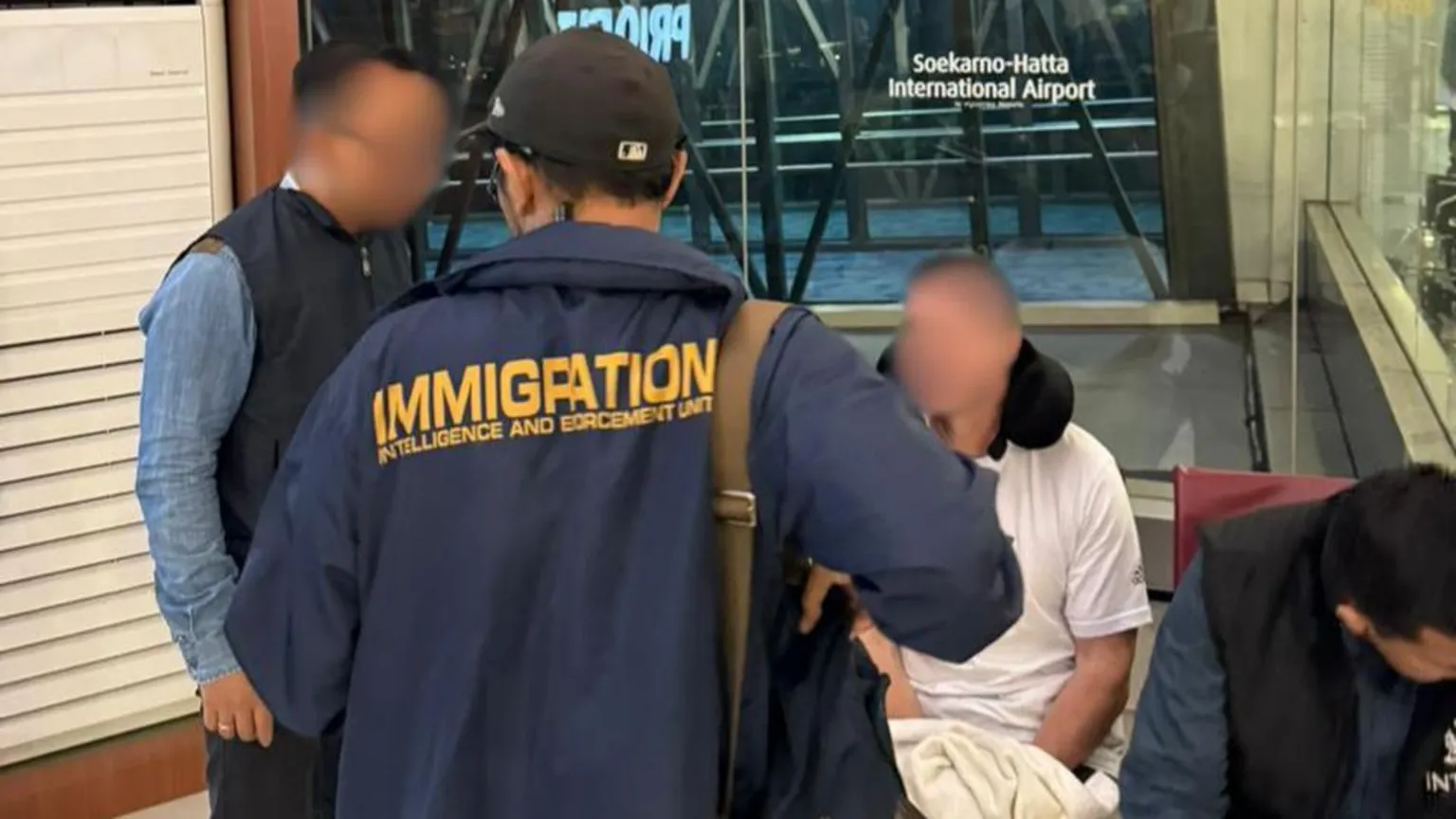 Ngurah Rai Immigration Office Two officials look at a man sitting down in an airport. It is Steven Lyons and he wears a black neck pillow and a white t-shirt. All of them have their faces blurred out