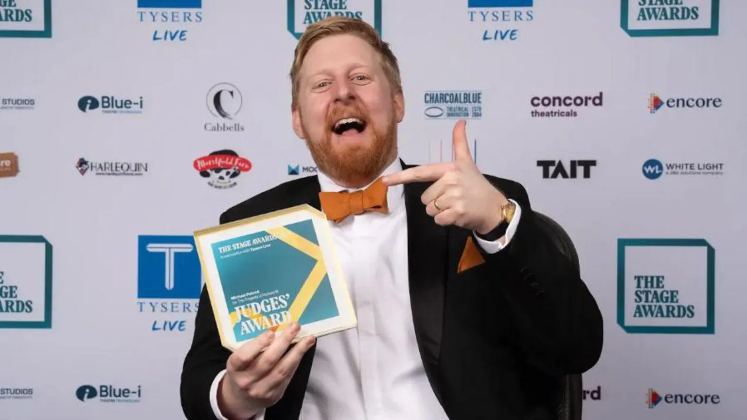 Alex Brenner Michael Campbell is wearing a black suit, white shirt and orange bow tie. He is seen holding the Judges' Award from the Stage Theatre Awards.