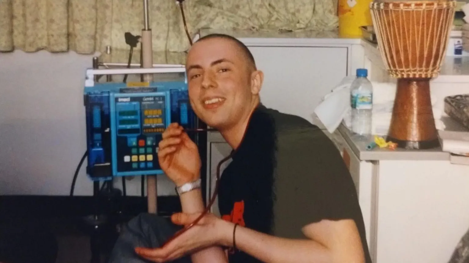 Jody White Jody White in hospital. He is wearing a black t-shirt and has a shaved head. He sits beside a hospital machine. He is smiling at the camera. 