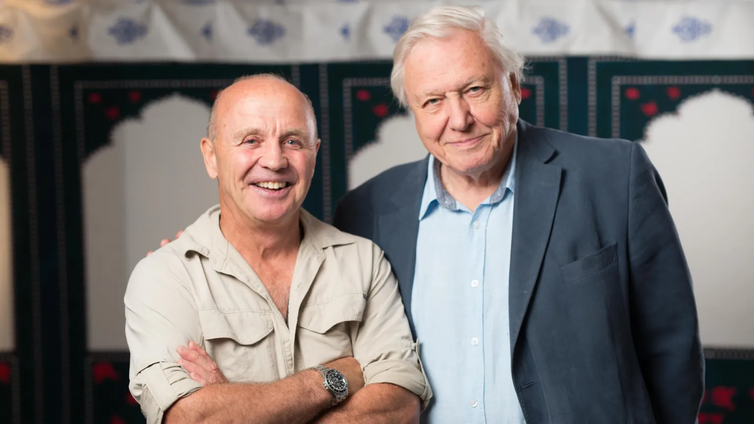  Doug Allan, left, wearing a beige shirt, with his arms folded across his chest. He is bald and is smiling at the camera. He is wearing a silver watch on his right wrist. Sir David Attenborough, right, wearing a grey suit jacket over a blue shirt. He is smiling and has white hair. He is standing in front of a white and green background.
