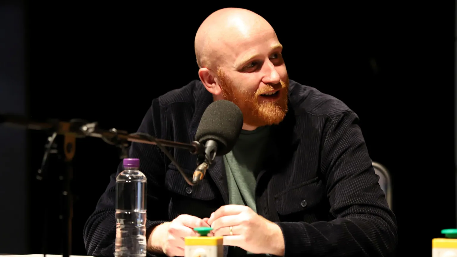 Ray Bradshaw - a bald man with a ginger beard, sitting behind a desk with a microphone in front of him. He is recording a panel comedy show for the radio.