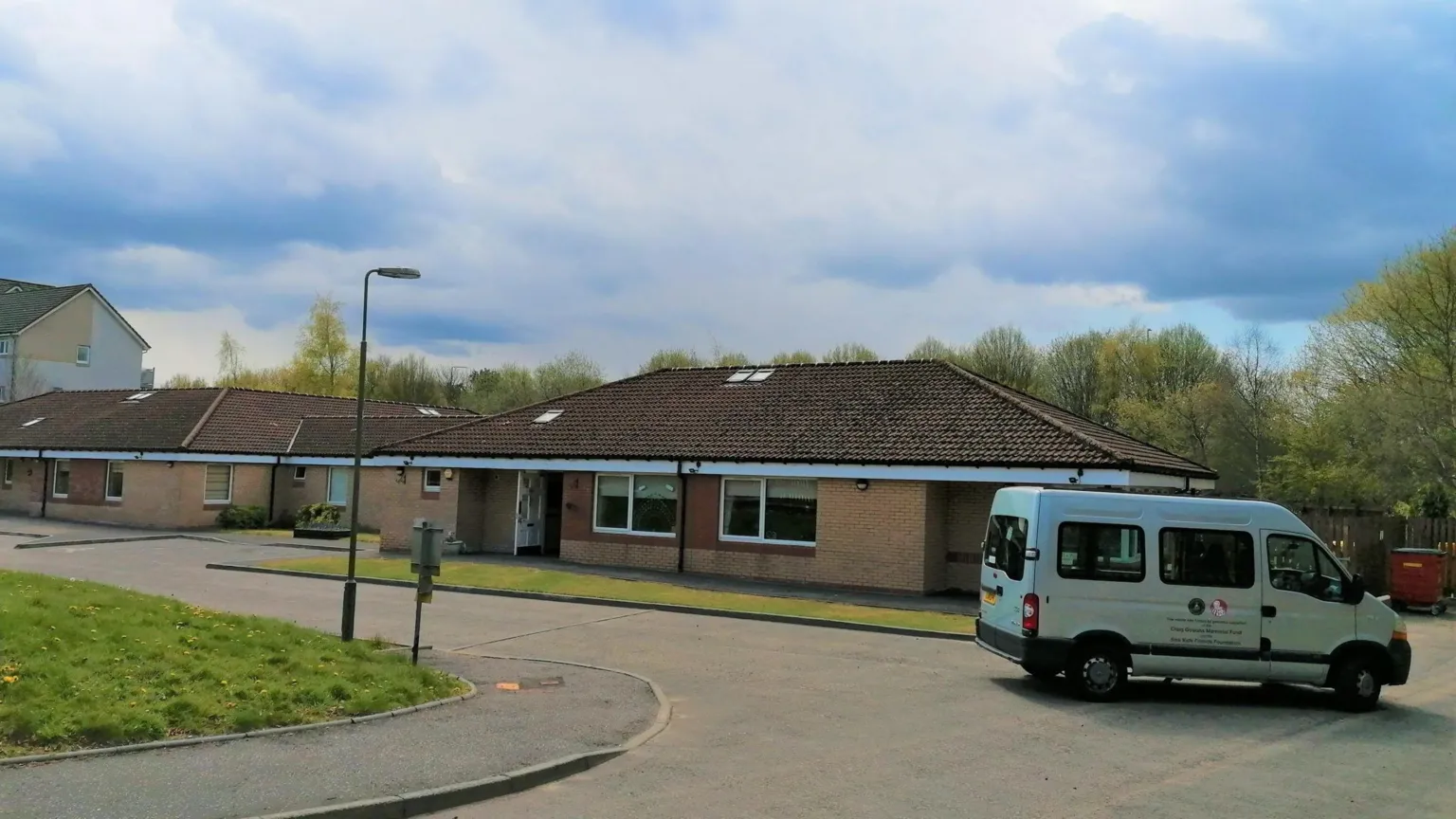NHS Lothian Exterior of a brown bungalow with a white van outside. It is the end of a street with a road that curves around it. There are blue, cloudy skies.