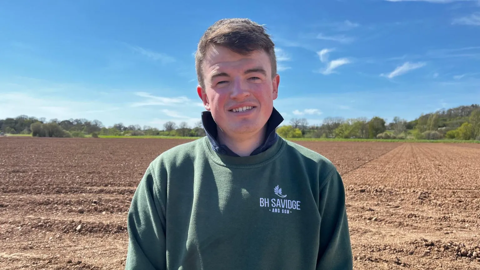 Ben Savidge stands on his farm looking at the camera. He wears a dark green jumper branded 'BH Savidge'.