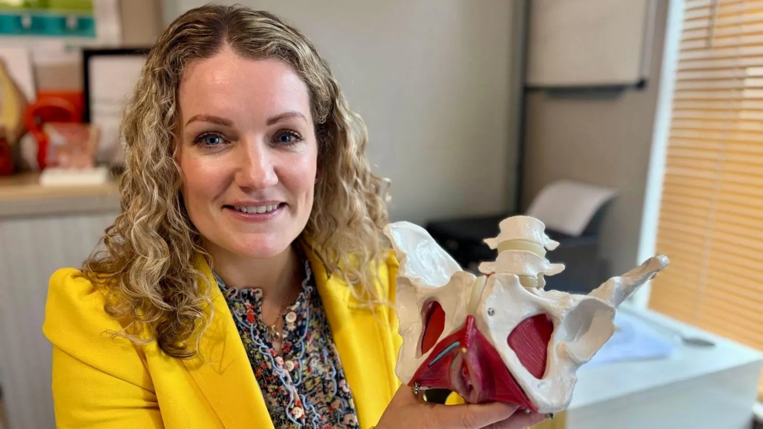 Gráinne is smiling at the camera. She is wearing a yellow blazer and a floral shirt. She has blond curly hair. She is holding a model of a pelvis. 