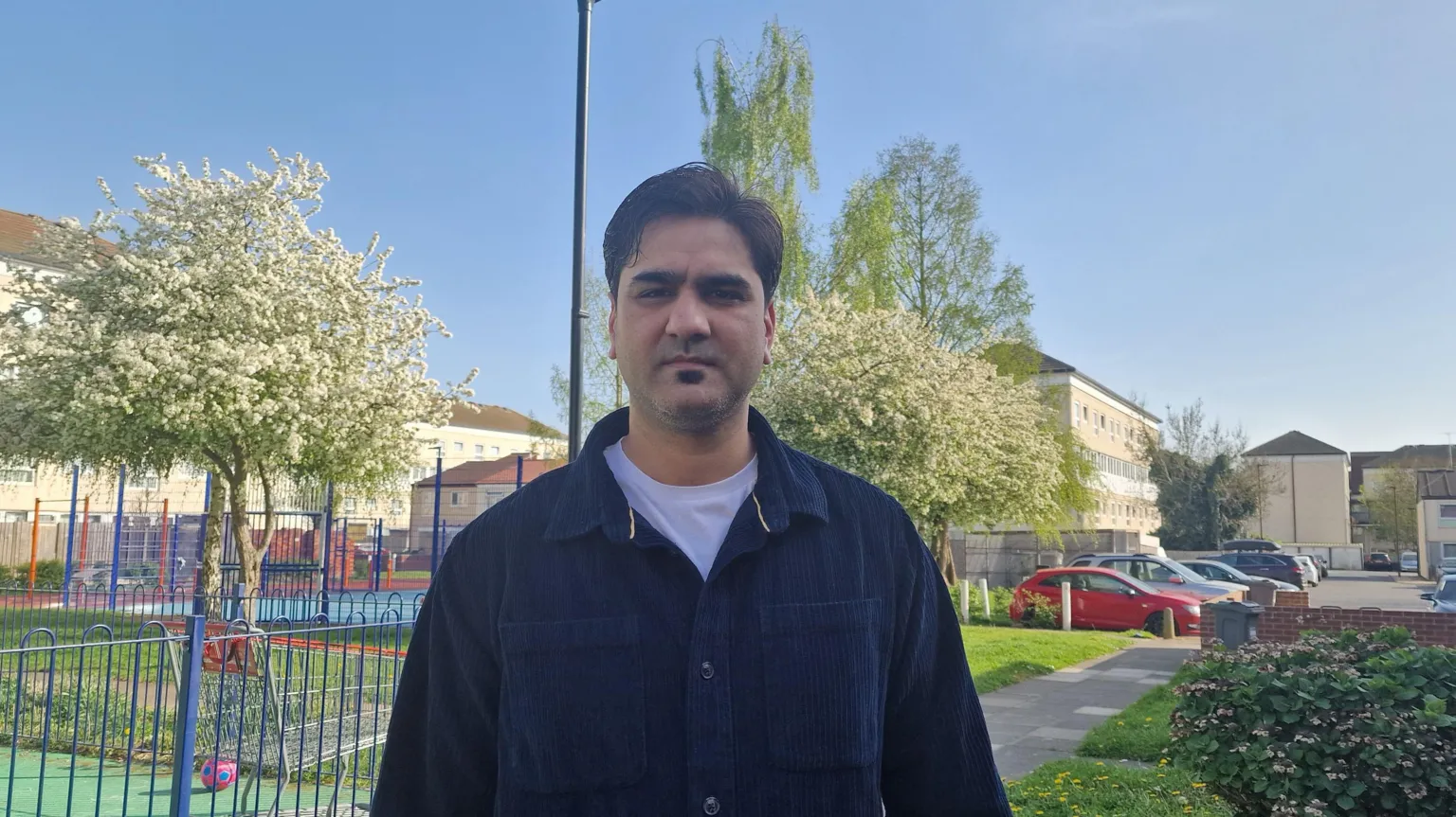 Handout A young Asian male wearing a white T-shirt under a dark blue shirt stands by a park in the centre of a housing estate