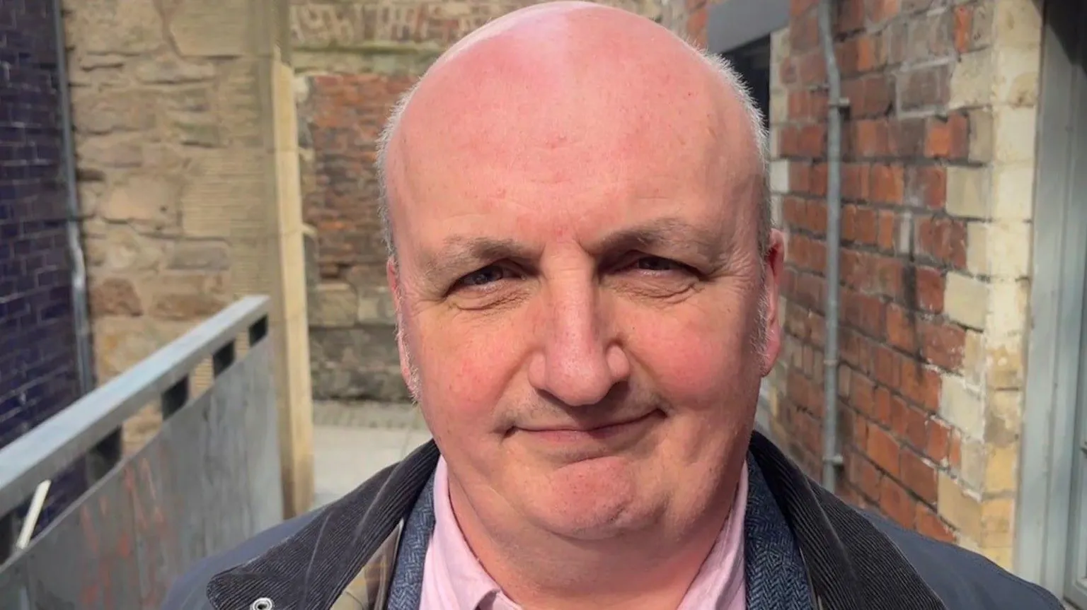 Martin Gallagher, a bald man, smiles without showing any teeth. He is wearing a pink shirt and a blue jacket