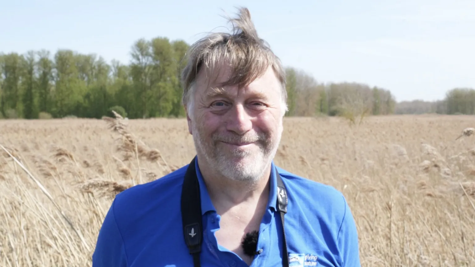 John Fairhall/BBC Dave Rogers stands in a wheat field and smiles at the camera. He has grey and brown hair, some of which rests over his forehead. He wears a blue top with a black binocular strap around his neck. 
