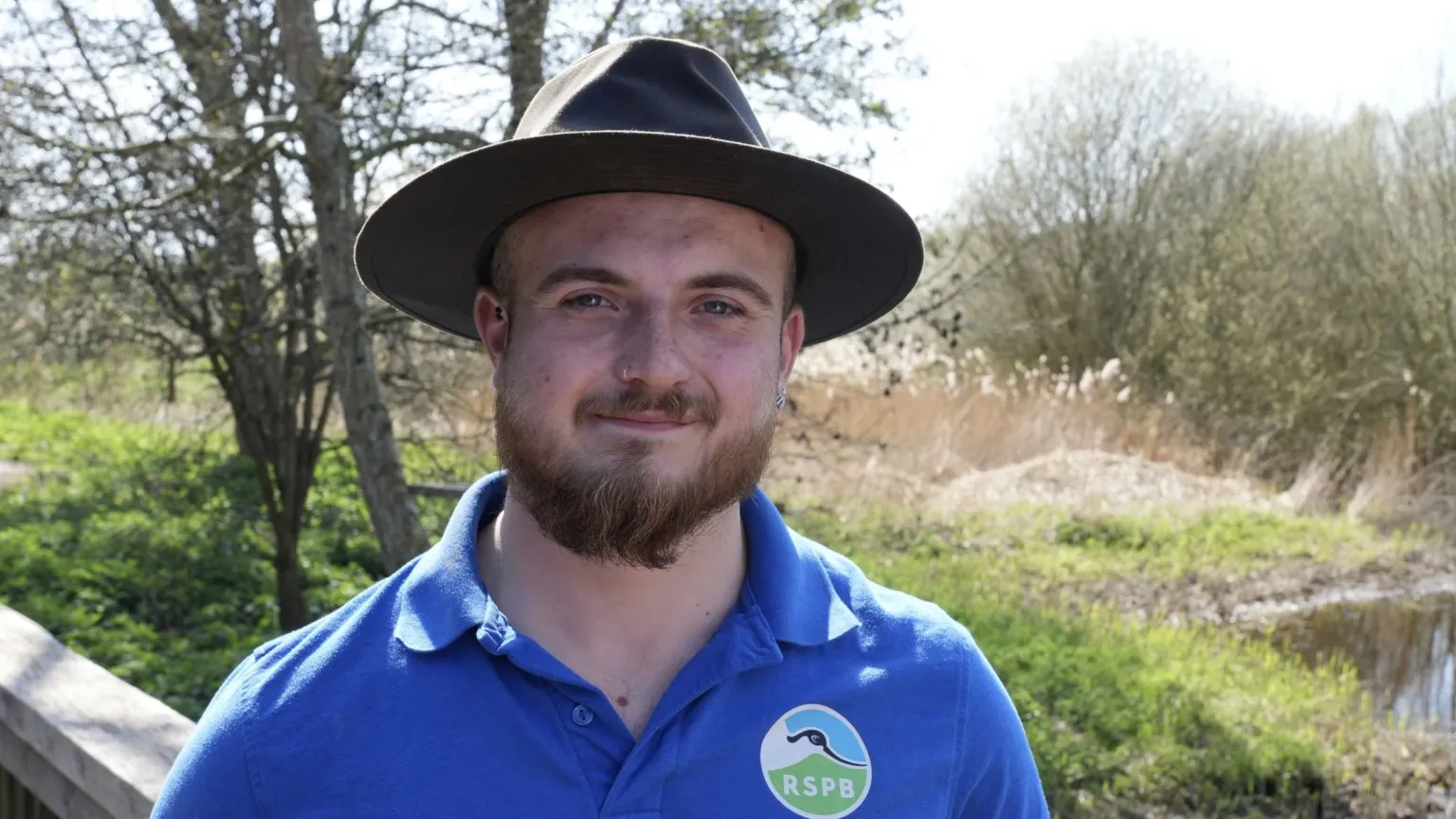 John Fairhall/BBC Haydn Fox stands in part of a nature reserve and smiles at the camera. He wears a large, dark coloured hat, and blue polo T-shirt. He has a thick beard.