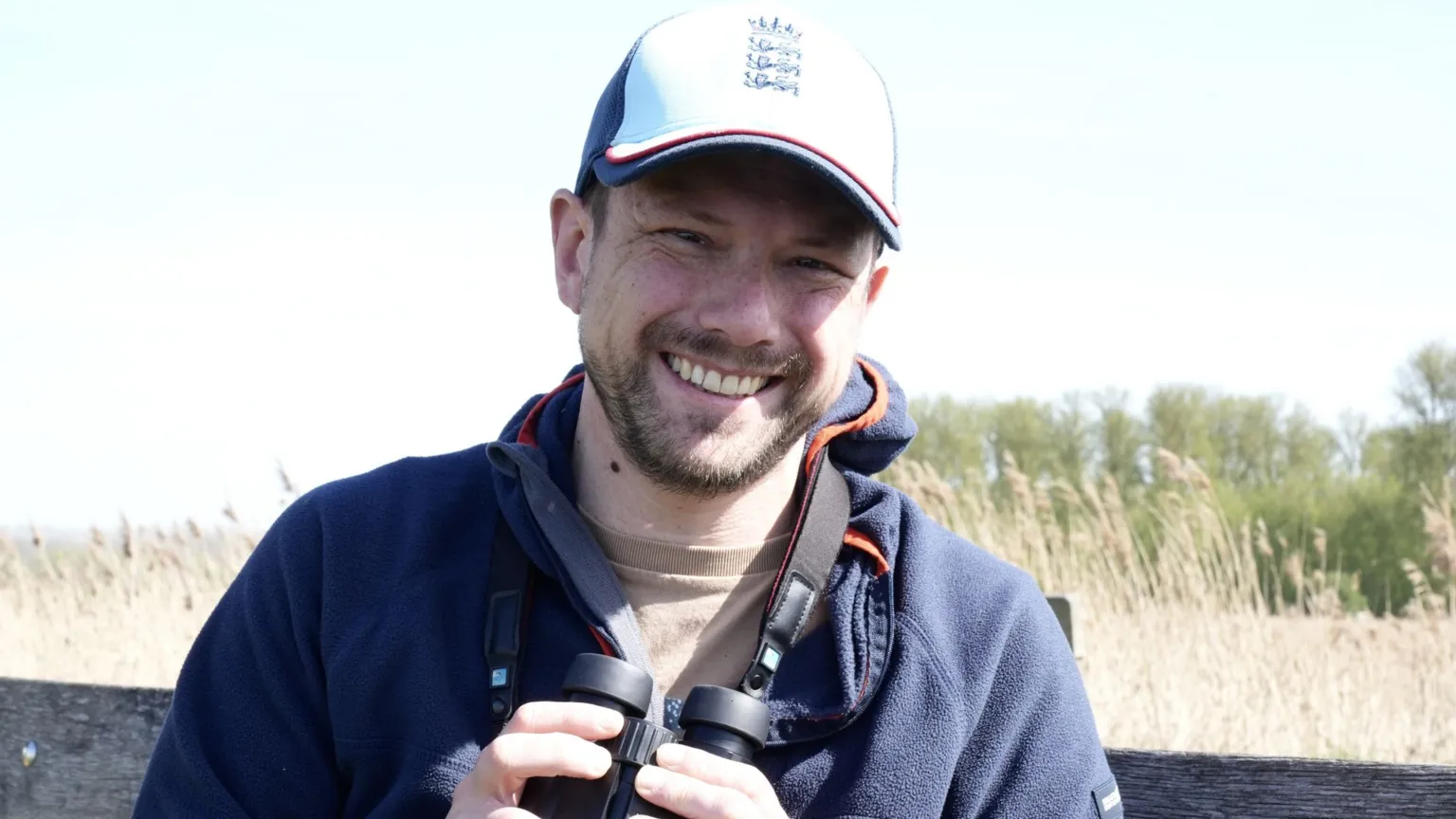 John Fairhall/BBC Kevin Middleton smiles at the camera as he holds a pair of binoculars while standing in a nature reserve. He wears a white cap with the England football logo on it. He is also wearing a blue fleece jumper and has some stubble on his face. 
