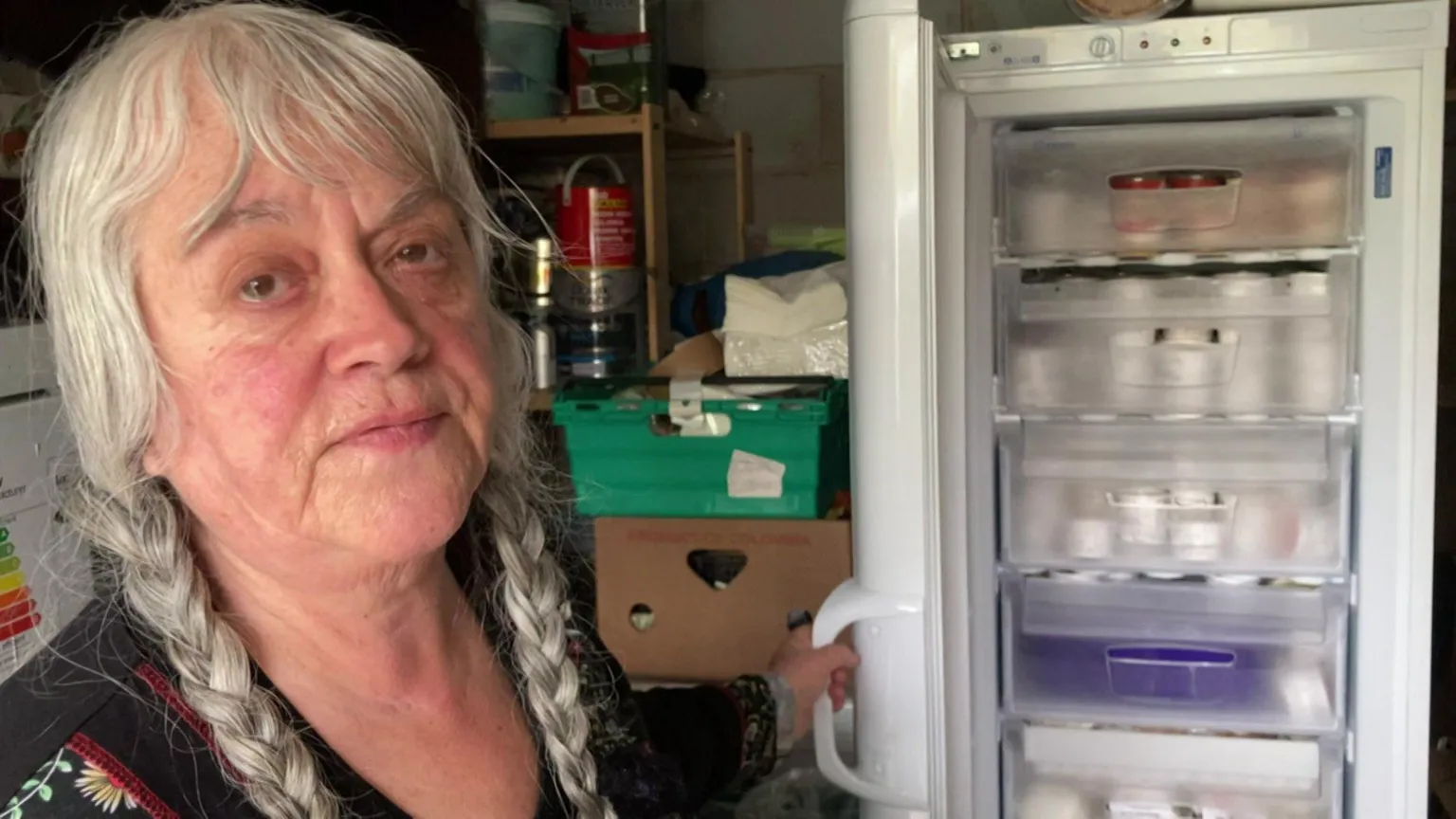 An older woman indoors, standing beside an open upright freezer. She has long grey hair styled into two braids and is wearing a dark top with a patterned neckline. The freezer is tall and white, with multiple transparent drawers visible inside. The drawers contain neatly stored food items, some in containers, giving the impression of organised storage. The surrounding environment looks like a garage. In the background, there are stacked boxes, shelves, and household supplies, suggesting a functional, everyday setting rather than a living room or kitchen. Overall, the image conveys a practical, domestic moment, focused on food storage and everyday life rather than a formal portrait.