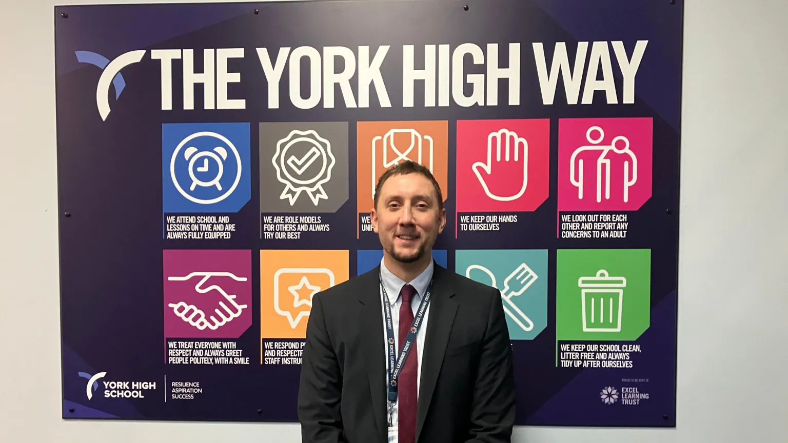 A man standing in front of a large wall display titled “THE YORK HIGH WAY.” The display features a set of colourful square icons, each representing a value or principle associated with York High School. These icons include imagery such as a rosette with a checkmark, a handshake, a hand raised, two people standing together, a star, a bin. They are each paired with short statements. The man is wearing a dark suit with a light shirt and a dark red tie. He also has a lanyard around their neck.